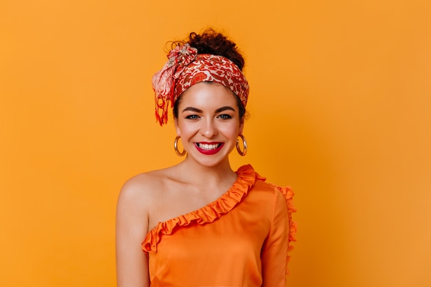 La chica joven de buen humor está sonriendo en el espacio naranja. Elegante dama de cabello oscuro con blusa naranja y bufanda en la cabeza mira a la cámara.