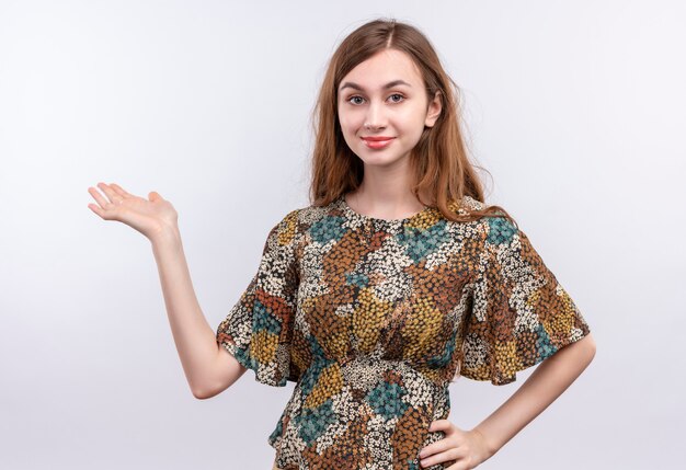 Chica joven con cabello largo vistiendo coloridos vestidos buscando confianza sonriendo presentando con el brazo de su mano