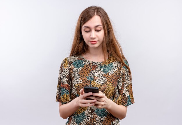 Chica joven con cabello largo vistiendo coloridos vestidos sosteniendo smartphone charlando con alguien con seria expresión de confianza en la cara