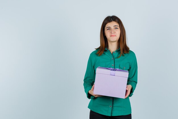 Chica joven con caja de regalo en ambas manos en blusa verde, pantalón negro y mirando disgustado. vista frontal.