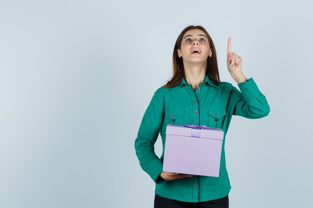 Chica joven con caja de regalo, apuntando hacia arriba con el dedo índice en blusa verde, pantalón negro y mirando feliz, vista frontal.