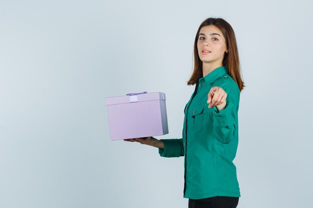 Chica joven con caja de regalo, apuntando a la cámara en blusa verde, pantalón negro y mirando alegre. vista frontal.