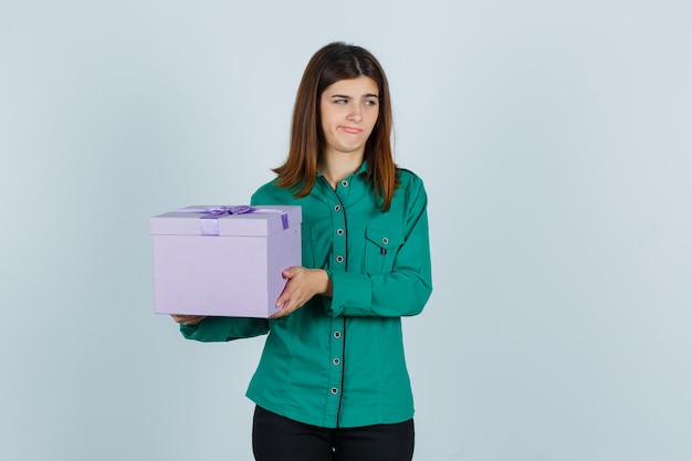 Chica joven con caja de regalo en blusa verde, pantalón negro y mirando disgustado. vista frontal.