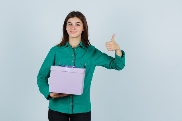 Chica joven con caja de regalo, mostrando el pulgar hacia arriba en blusa verde, pantalón negro y mirando alegre, vista frontal.