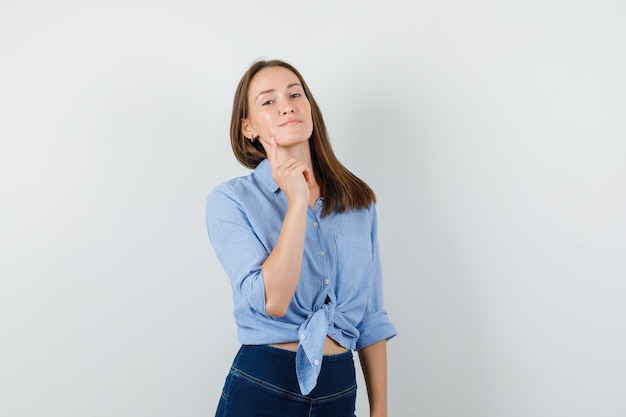 Chica joven con camisa azul, pantalones sosteniendo el dedo en la barbilla y mirando confiado