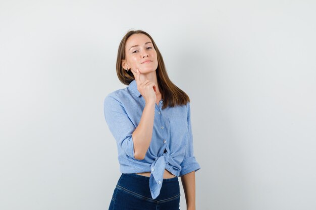 Chica joven con camisa azul, pantalones sosteniendo el dedo en la barbilla y mirando confiado