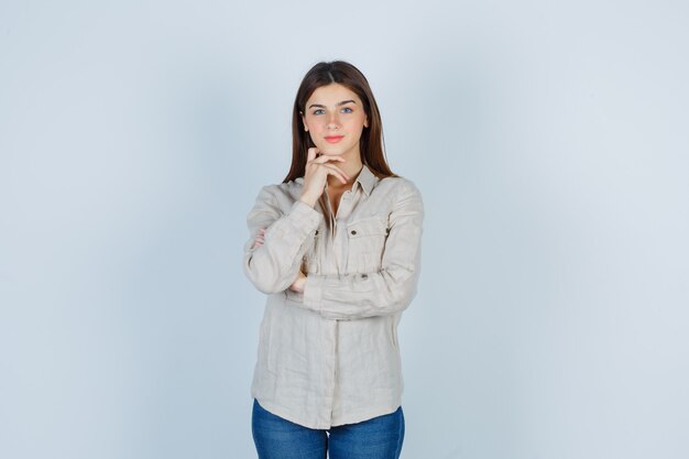 Chica joven en camisa beige, jeans apoyando la barbilla en la mano y mirando alegre, vista frontal.