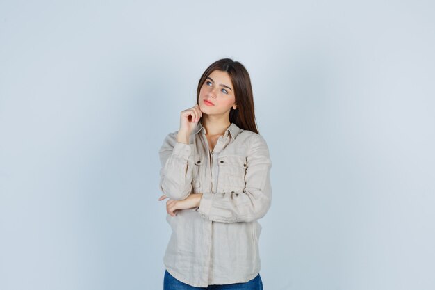 Chica joven con camisa beige, jeans apoyando la barbilla en la mano, pensando en algo y mirando pensativa, vista frontal.