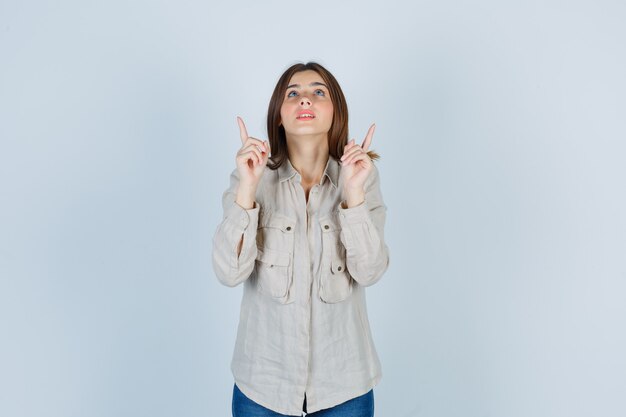 Chica joven en camisa beige, jeans apuntando hacia arriba con los dedos índices y mirando enfocado, vista frontal.