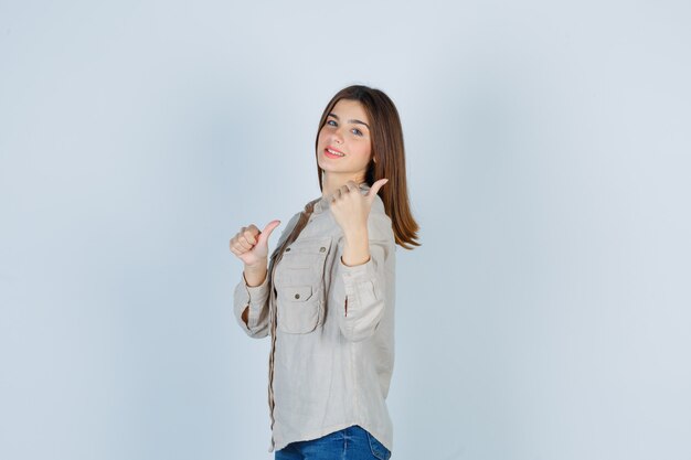Chica joven en camisa beige, jeans apuntando a un lado con los pulgares y mirando feliz, vista frontal.