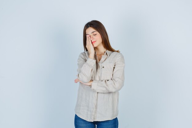 Chica joven en camisa beige, jeans cubriendo los ojos con la mano y con aspecto cansado, vista frontal.