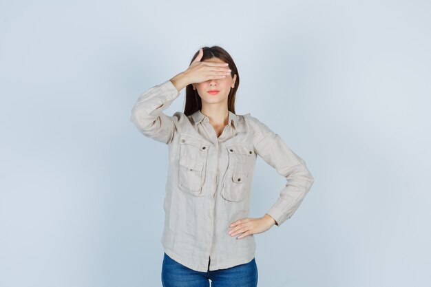 Chica joven en camisa beige, jeans cubriendo los ojos con la mano, con la mano en la cadera y mirando seria, vista frontal.