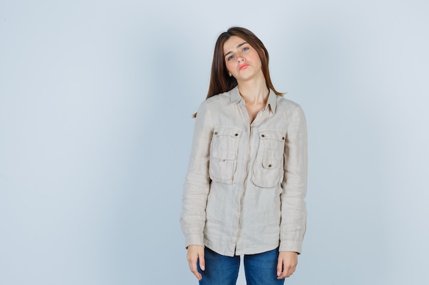 Chica joven en camisa beige, jeans doblando la cabeza mientras posa y luce linda, vista frontal.