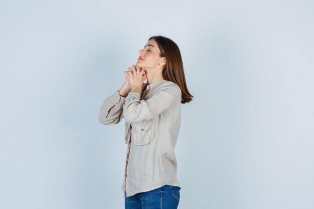 Chica joven en camisa beige, jeans estrechando las manos en posición de oración y mirando esperanzado, vista frontal.