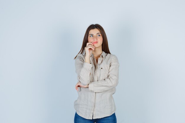 Chica joven en camisa beige, jeans con la mano cerca de la boca, pensando en algo y mirando pensativa, vista frontal.