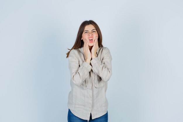 Chica joven en camisa beige, jeans con las manos cerca de la boca, haciendo muecas y luciendo linda, vista frontal.