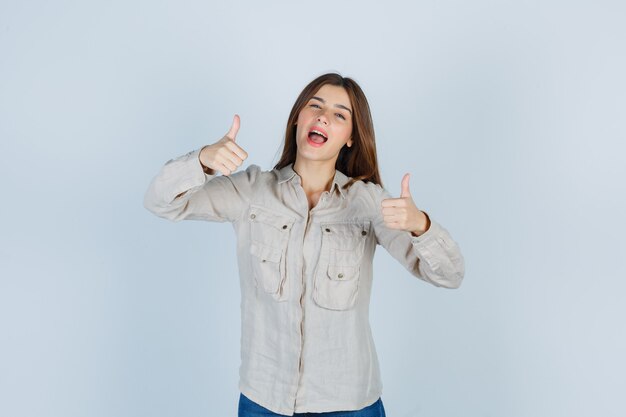 Chica joven en camisa beige, jeans mostrando doble pulgar hacia arriba y mirando alegre, vista frontal.