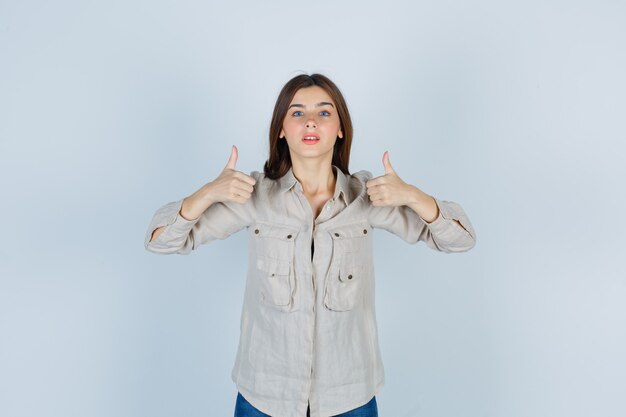 Chica joven en camisa beige, jeans mostrando doble pulgar hacia arriba y mirando confiado, vista frontal.