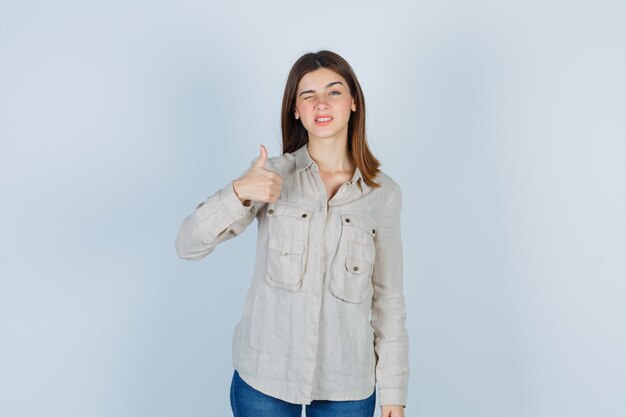 Chica joven en camisa beige, jeans mostrando el pulgar hacia arriba, guiñando un ojo y luciendo linda, vista frontal.