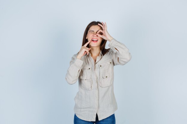 Chica joven con camisa beige, jeans mostrando signo ok en el ojo, poniendo el dedo índice cerca de la boca y luciendo linda vista frontal.