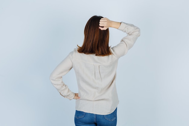 Chica joven con camisa beige, jeans sosteniendo una mano detrás de la cabeza, otra mano en la cadera y luciendo linda vista frontal.
