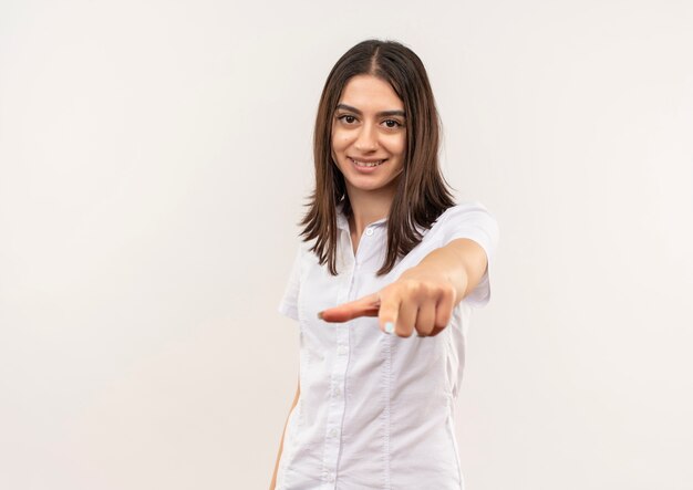 Chica joven con camisa blanca apuntando con el dedo hacia el frente sonriendo confiado de pie sobre la pared blanca