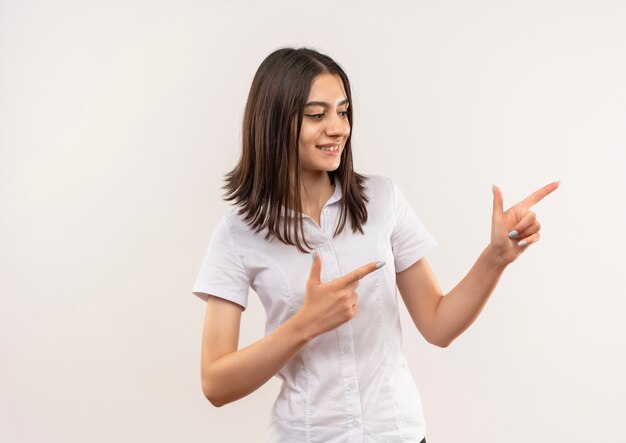 Chica joven con camisa blanca apuntando con el dedo índice hacia el lado sonriendo de pie sobre la pared blanca