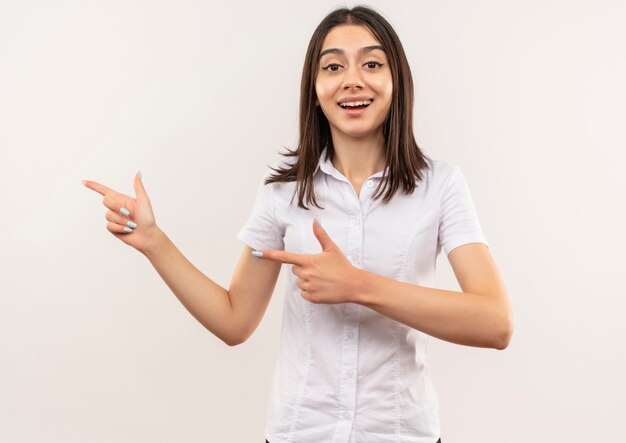 Chica joven con camisa blanca apuntando con el dedo índice hacia el lado sonriendo de pie sobre la pared blanca