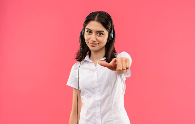 Chica joven con camisa blanca y auriculares, apuntando con el dedo índice hacia el frente mirando confiado de pie sobre la pared rosa