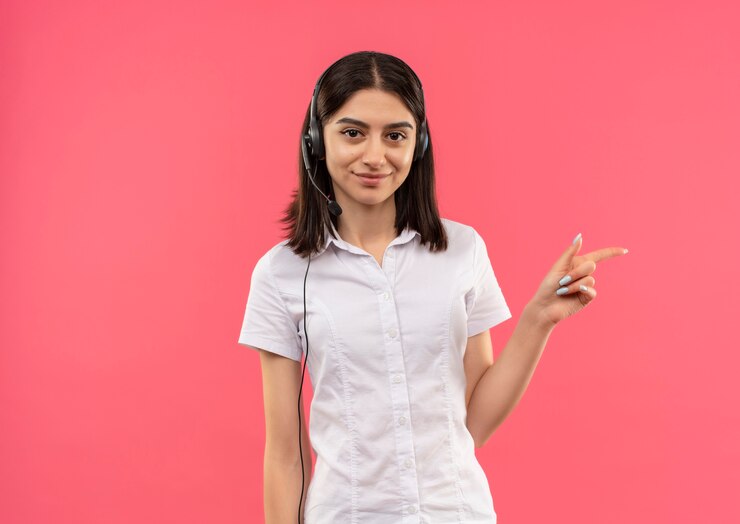Foto gratuito chica joven con camisa blanca y auriculares, apuntando con el dedo hacia el lado sonriendo de pie sobre la pared rosa
