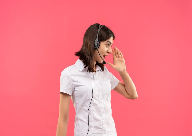 Chica joven con camisa blanca y auriculares, gritando o llamando a alguien con la mano cerca de la boca de pie sobre una pared rosa