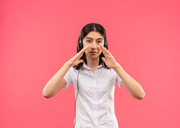 Chica joven con camisa blanca y auriculares, gritando o llamando a alguien con las manos cerca de la boca de pie sobre una pared rosa