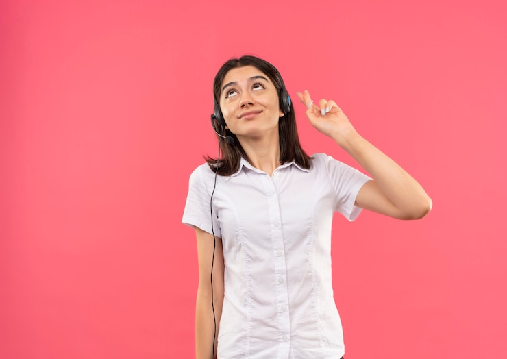 Foto gratuito chica joven con camisa blanca y auriculares, haciendo un deseo deseable cruzar los dedos de pie feliz y positivo sobre la pared rosa
