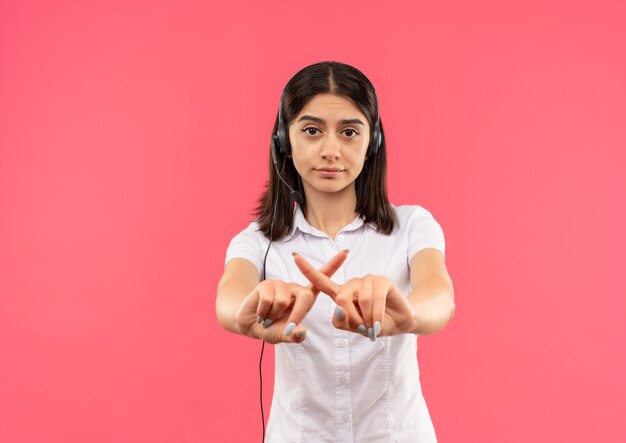 Chica joven con camisa blanca y auriculares, haciendo que deje de cantar cruzando los dedos índices de pie sobre la pared rosa