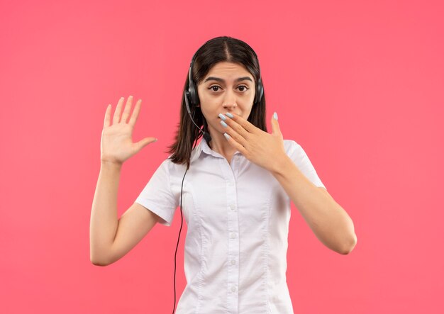 Chica joven con camisa blanca y auriculares, mirando al frente asombrado y sorprendido cubriendo la boca con la mano de pie sobre la pared rosa