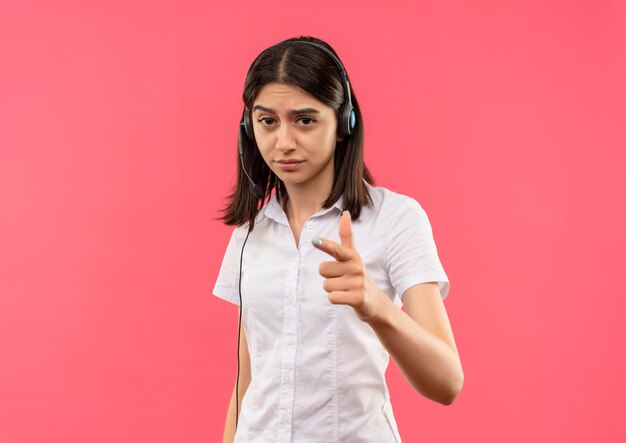 Chica joven con camisa blanca y auriculares, mirando al frente con cara seria apuntando con el dedo hacia el frente parado sobre pared rosa