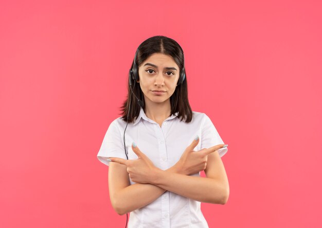 Chica joven en camisa blanca y auriculares, mirando al frente con cara seria apuntando con los dedos a los lados de pie sobre la pared rosa