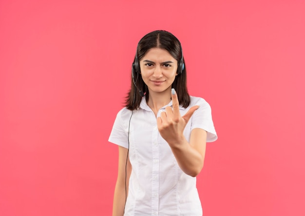 Chica joven con camisa blanca y auriculares, mirando al frente disgustado mostrando el dedo índice de pie sobre la pared rosa