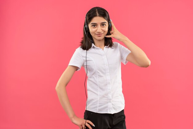 Chica joven con camisa blanca y auriculares, mirando al frente haciendo gesto de llamarme sonriendo de pie sobre la pared rosa