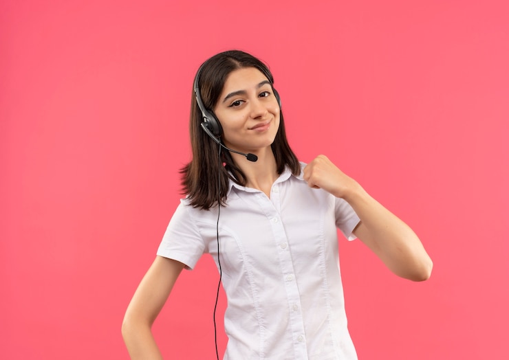 Foto gratuito chica joven en camisa blanca y auriculares, mirando al frente sonriendo apretando el puño, concepto ganador de pie sobre la pared rosa
