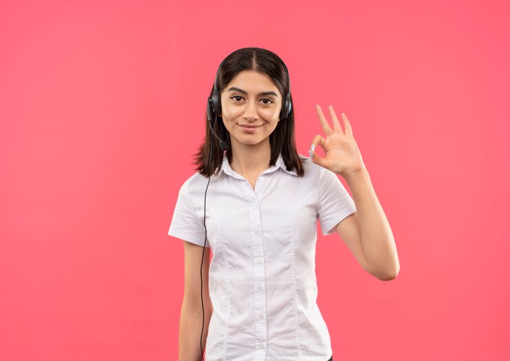 Foto gratuito chica joven con camisa blanca y auriculares, mirando al frente sonriendo mostrando signo ok de pie sobre la pared rosa