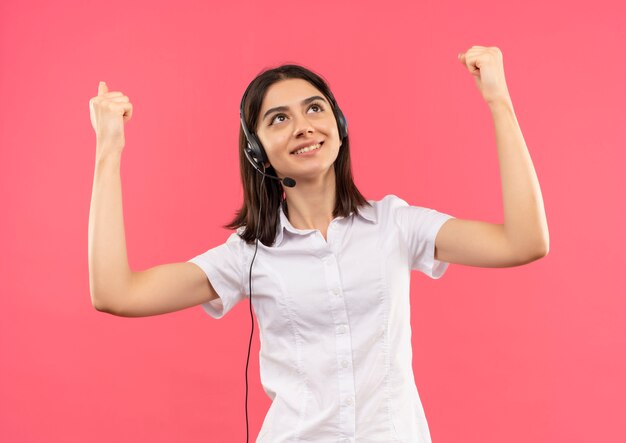 Chica joven con camisa blanca y auriculares, mirando feliz y emocionado sonriendo apretando los puños, concepto ganador de pie sobre la pared rosa