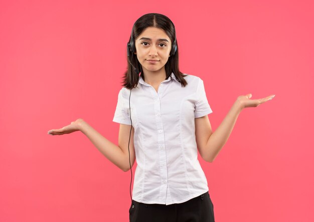 Chica joven con camisa blanca y auriculares, mirando hacia el frente confundido extendiendo los brazos a los lados parados sobre la pared rosa