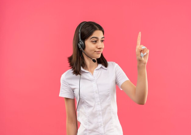 Chica joven con camisa blanca y auriculares, mirando a un lado mostrando el dedo índice de pie sobre la pared rosa