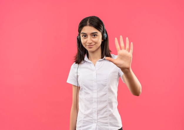 Chica joven con camisa blanca y auriculares, mostrando y señalando con los dedos el número cinco sonriendo de pie sobre la pared rosa
