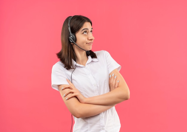 Chica joven con camisa blanca y auriculares, poniendo los ojos en blanco mirando disgustado parado sobre la pared rosa