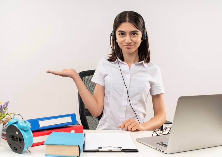 Foto gratuito chica joven con camisa blanca y auriculares, presentando algo con el brazo de su mano sonriendo sentado en la mesa con carpetas y portátil sobre pared blanca