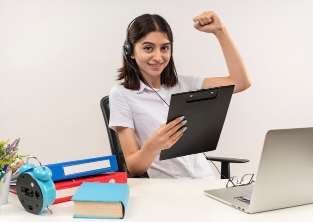 Chica joven con camisa blanca y auriculares, sosteniendo el portapapeles apretando el puño feliz y positivo sentado en la mesa con carpetas y portátil sobre pared blanca