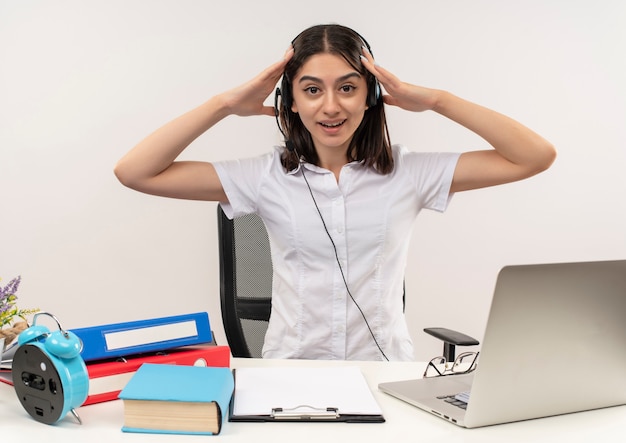 Chica joven con camisa blanca y auriculares, sosteniendo su cabeza con las manos mirando confundido sentado en la mesa con carpetas y computadora portátil sobre pared blanca