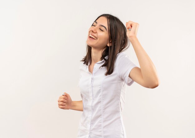 Chica joven en camisa blanca feliz y positiva con los ojos cerrados de pie sobre la pared blanca